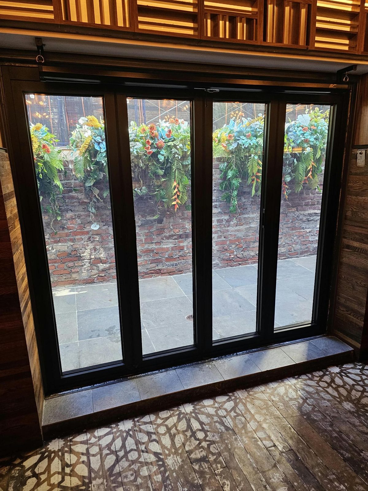Four-panel black aluminum bifold door seen from the restaurant dining room at 107 1st Avenue, East Village, Manhattan, looking onto the planted brick patio wall