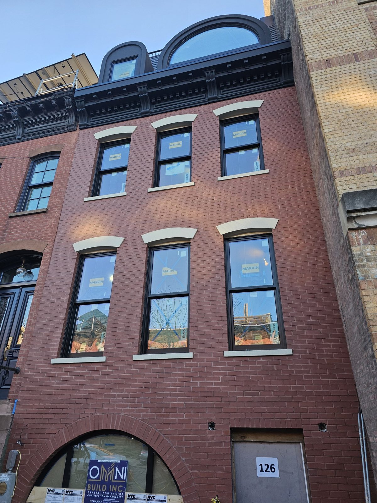Brick townhouse front at 126 Washington Avenue, Clinton Hill, Brooklyn, with six dark Marvin double-hung windows under curved stone lintels and an arched aluminium dormer window above the black cornice