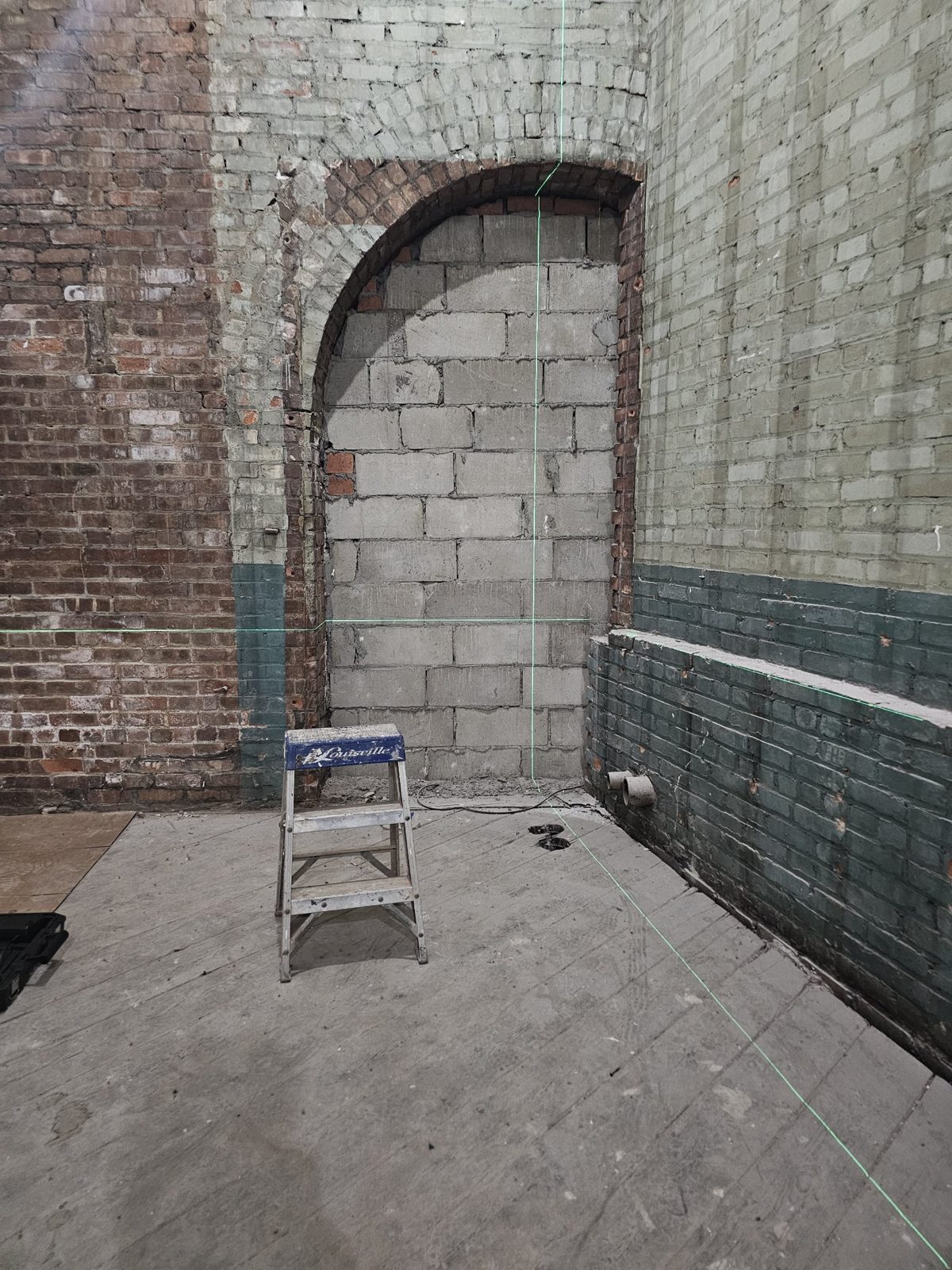Before: an arched brick opening at 185 Van Dyke Street, Red Hook, closed up with concrete block, seen from inside with green laser level lines projected on the wall
