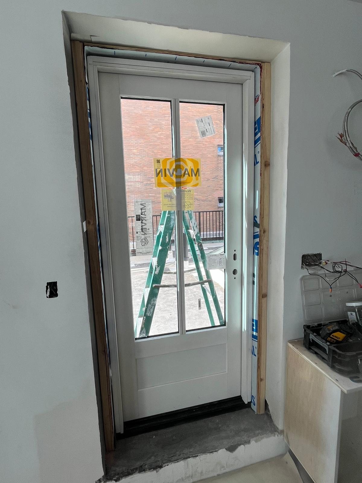 White Marvin outswing terrace door with two vertical lites over a raised panel, seen from inside the penthouse during installation, terrace railing beyond
