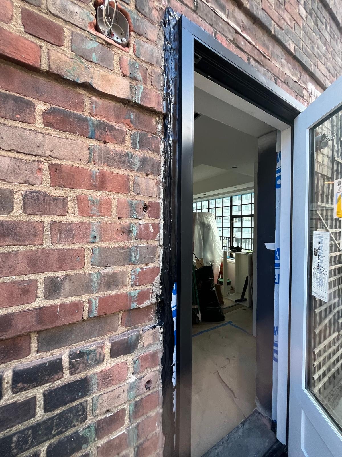 Exterior of the Marvin terrace door from the penthouse terrace: dark-clad frame, membrane flashing into the brick, door open to the room with steel windows beyond
