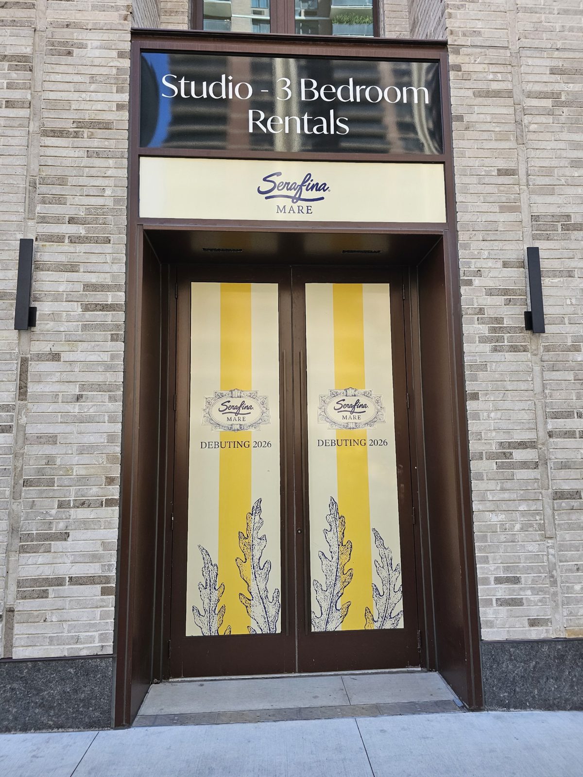 Double-leaf aluminium entry doors in a dark bronze frame set in a brick opening at 300 East 50th Street, Manhattan, with a sign panel in the transom above