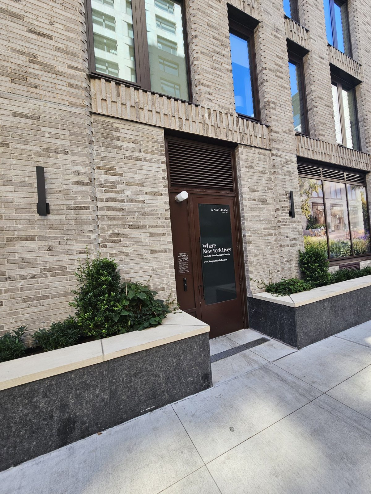 Single-leaf entry door in a dark bronze aluminium frame set in the brick at 300 East 50th Street, Manhattan, with a glazed curtain wall bay and planters beside it