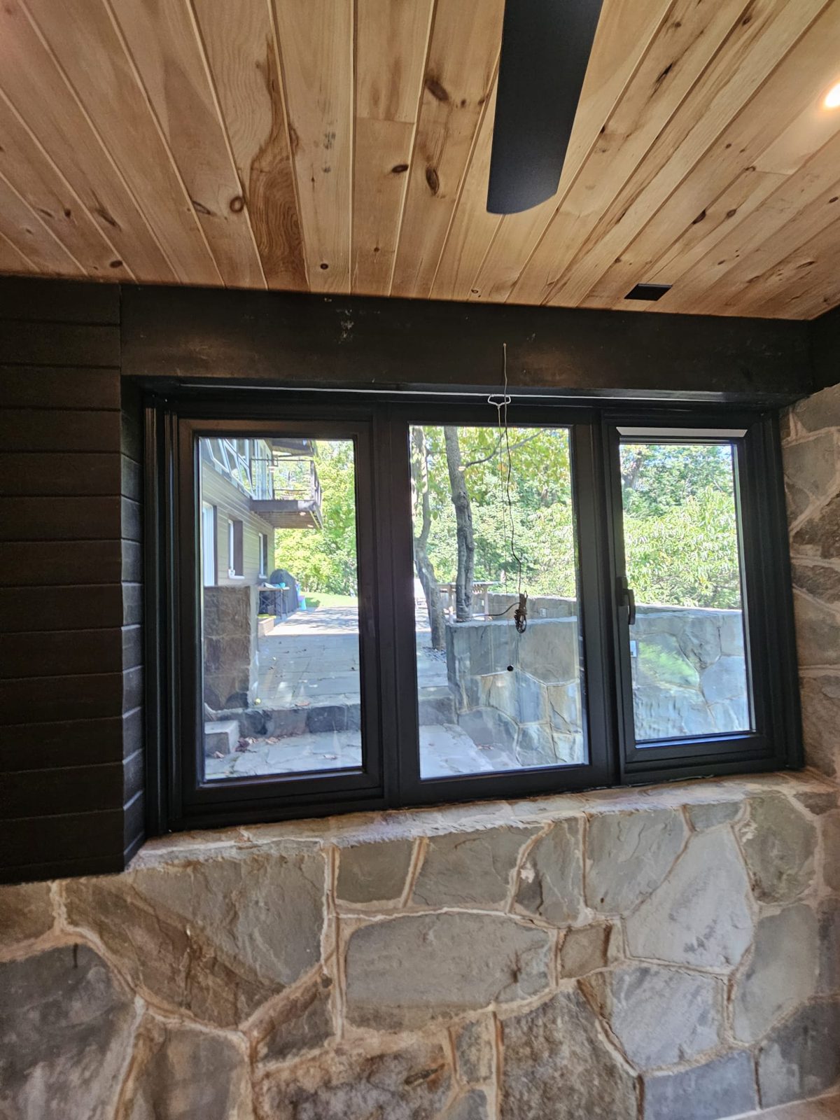 Three-lite black aluminium casement window set in a fieldstone wall under a wood-plank ceiling, looking onto a bluestone terrace, sunroom near Morristown NJ