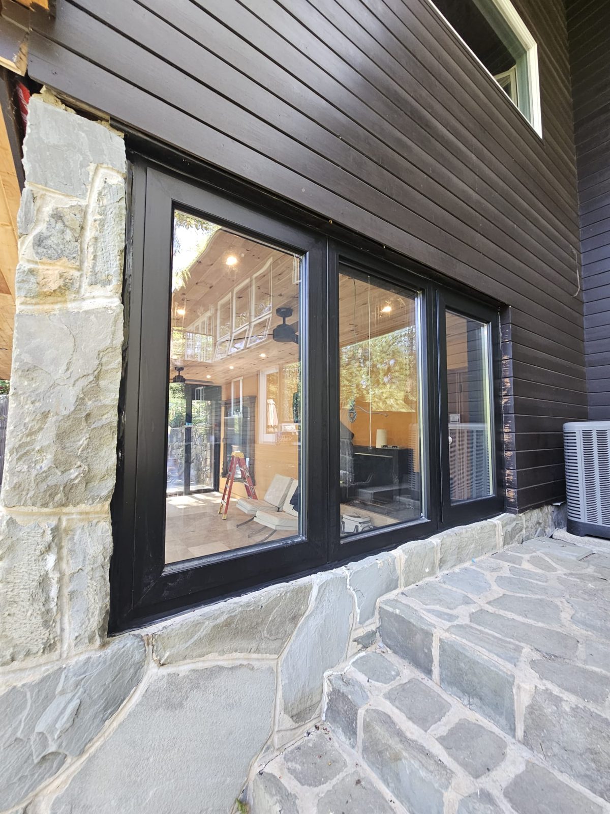 Exterior of the black aluminium three-lite casement window between fieldstone and dark board siding, with the sunroom interior visible through the glass