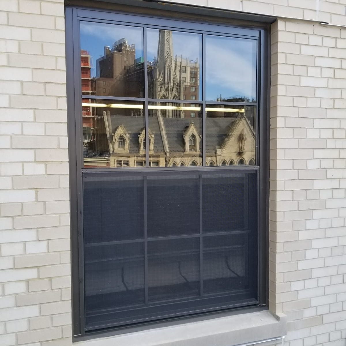 Dark bronze double-hung replacement window with divided lites and a half insect screen in a light brick wall at a U.S. Post Office near Webster Hall, Manhattan