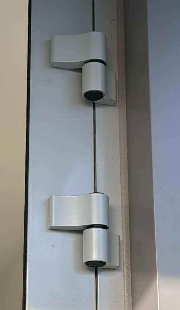 Two silver surface-mounted European door hinges fixed to the face of an aluminum door frame and door leaf