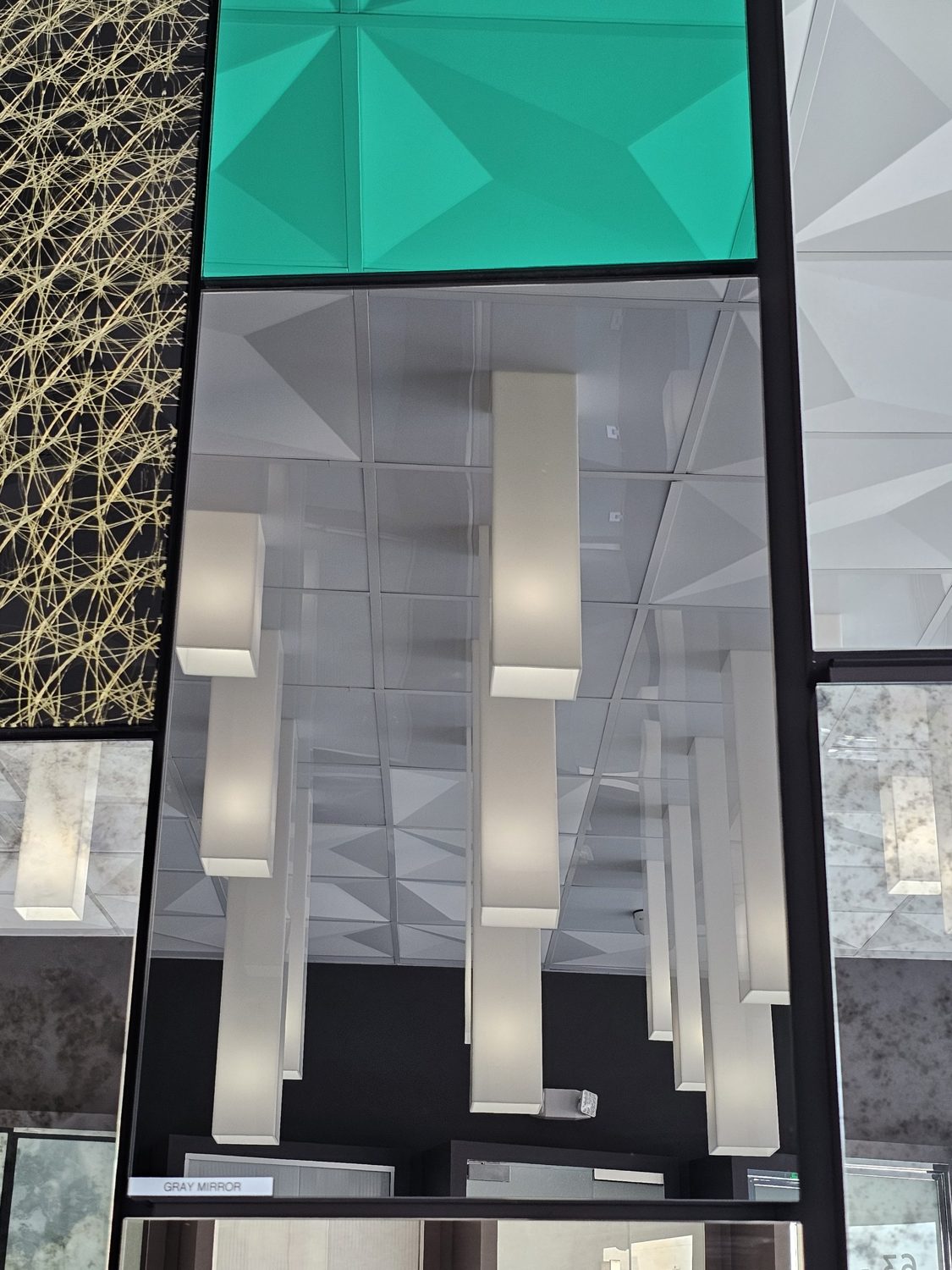 Gray mirror (smoke mirror) sample panel on a showroom display wall, reflecting ceiling lights in a darker, cooler tone, beside antique mirror and green back-painted glass samples