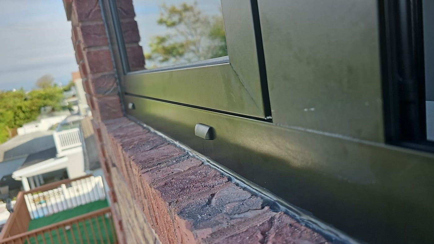 Weep hole covers on the outside face of a black aluminum window frame, above a sealed joint to a brick sill